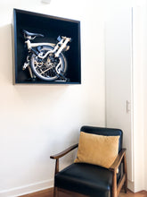 Load image into Gallery viewer, Brompton bicycle storage box mounted on wall with chair in the image to give the storage box scale. Custom designed hardwood wall storage including mitered corners, recessed bevel front edge, and black satin finish.