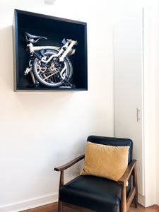 Brompton bicycle storage box mounted on wall with chair in the image to give the storage box scale. Custom designed hardwood wall storage including mitered corners, recessed bevel front edge, and black satin finish.
